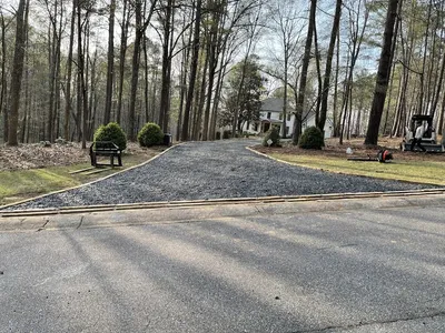 New gravel driveway installation leading to a white residential home surrounded by tall pine trees